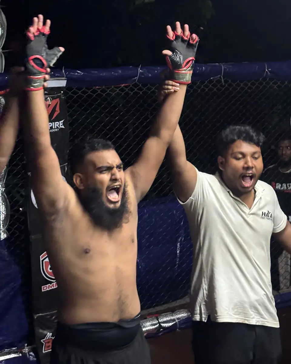Meer Estiyak with both arms raised after a win, the referee beside him in the cage